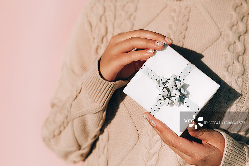 匿名女人穿着温暖的针织毛衣，在粉红色背景的节日庆祝活动中展示带丝带的时尚礼物。新年庆祝概念图片素材