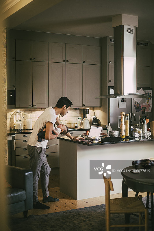 父亲和蹒跚学步的儿子在厨房里用笔记本电脑工作图片素材