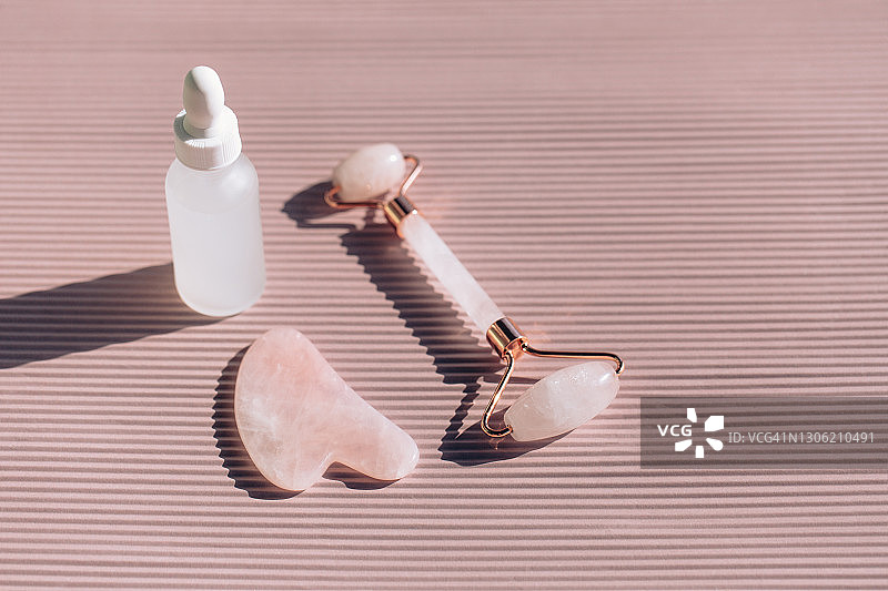 玫瑰石英滚轮和刮痧板靠近条纹粉红色背景上的白色磨砂玻璃瓶。按摩器与油或乳液一起用于抗衰老。阳光使瓶子产生阴影和照明反射。扁平风格图片素材