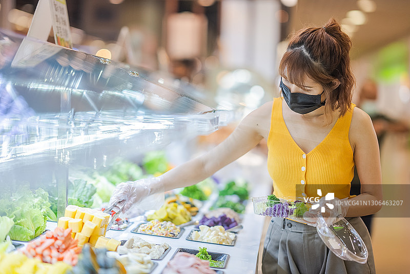 美丽的亚洲妇女戴着防护口罩，在自助服务中挑选食物，亚洲女性购买沙拉外卖食品，因冠状病毒隔离结束后新的正常生活方式图片素材