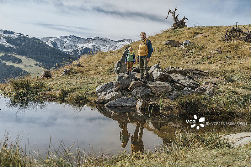 年长的男人和儿子站在奥地利萨尔茨堡地区莱奥冈山脉池塘边的岩石上，以天空为背景图片素材
