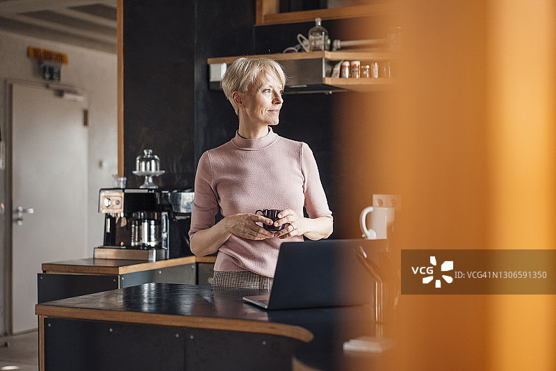 微笑的女商人拿着咖啡杯在家庭办公室厨房里看向远方图片素材