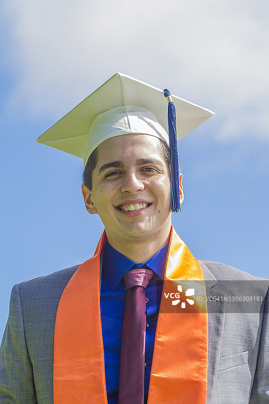在美国毕业典礼上，戴着帽子和礼袍的快乐英俊拉丁裔男学生肖像图片素材