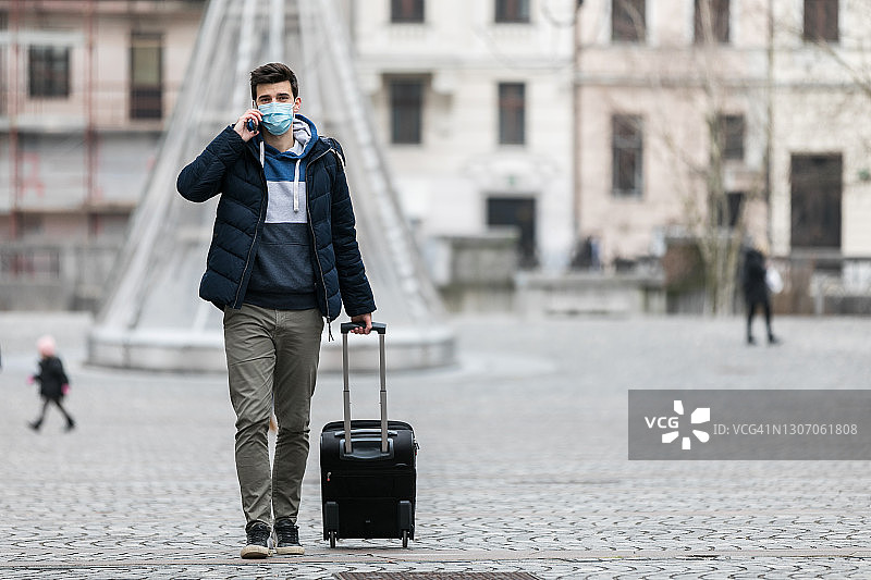 疫情期间戴一次性医用口罩的男人在路边叫出租车，拿着行李箱图片素材