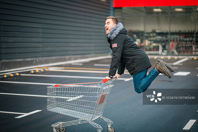 微笑的年轻男人在商店前跳跃，推着购物车图片素材