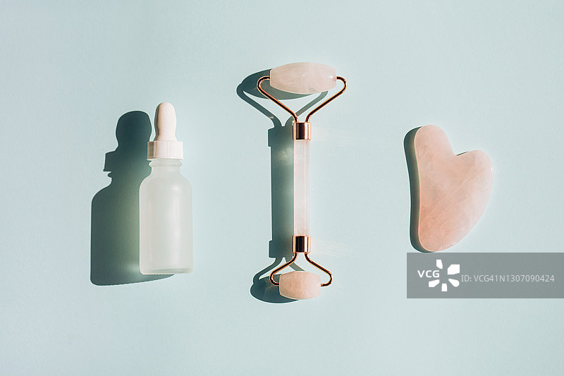 玫瑰石英滚轮和刮痧板，在粉蓝色背景上白色磨砂玻璃瓶面部精华液旁，按摩器与精油或保湿霜一起用于抗衰老，健康水疗和身体护理概念，具有复制空间的平面风格图片素材