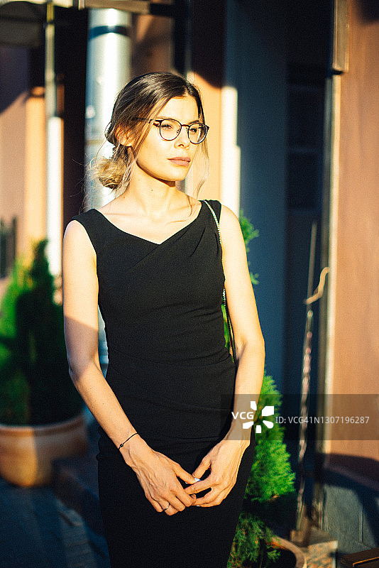 在阳光明媚的城市里，身穿黑色连衣裙和戴眼镜的年轻女人图片素材