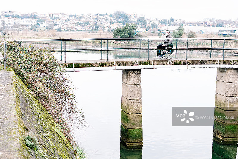 戴着口罩，坐在轮椅上站在河上桥中央的残疾男子图片素材