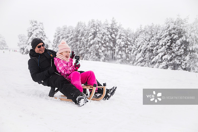 父女在雪橇上使用迷你相机时欢笑图片素材