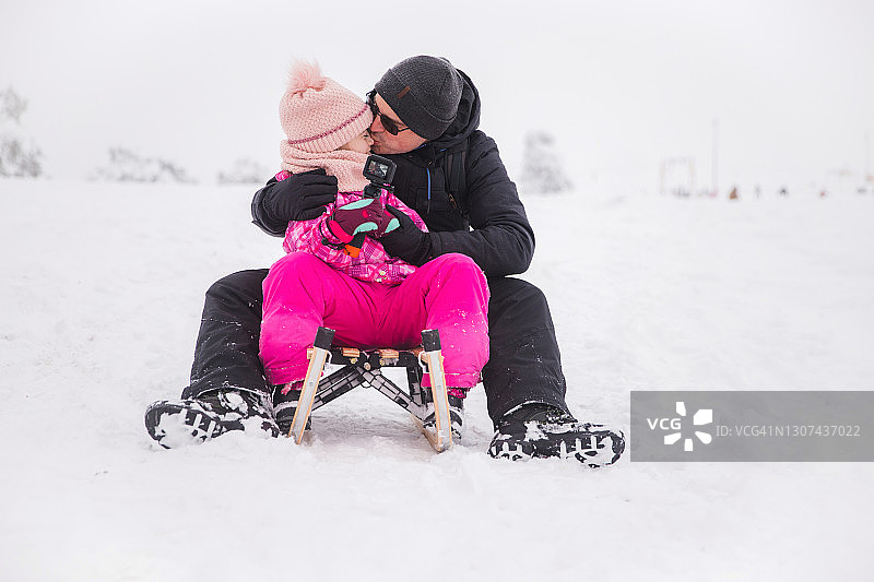 当父女两人在玩雪橇并通过GoPro记录乐趣时，男人亲吻他的女儿图片素材