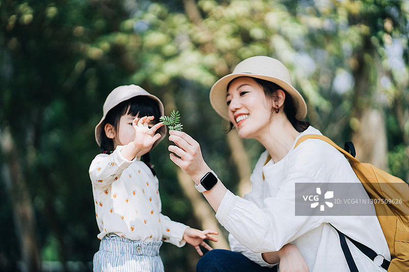年轻的亚洲母亲和小女儿在春天户外共度时光。小女儿看着妈妈拿着的树叶,探索大自然。快乐幸福的家庭时光,健康的家庭生活方式图片素材