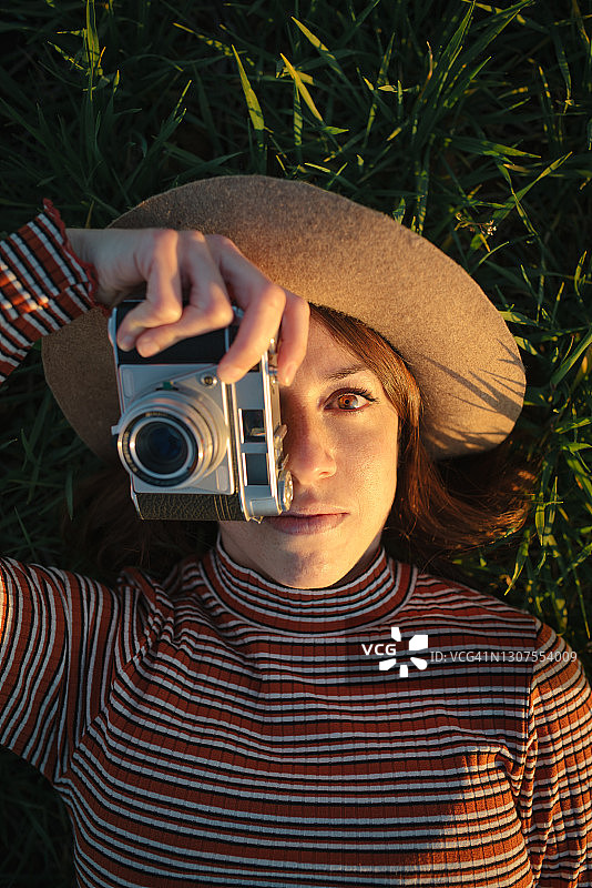 日落时分，身穿复古条纹裙的年轻白人女性在草地上用旧式胶片相机拍照特写图片素材