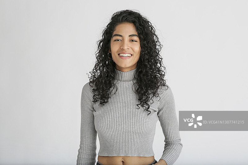 一位有着卷发的拉丁裔千禧一代女性，站在工作室的白色背景前，穿着灰色露脐上衣和蓝色牛仔裤的画像图片素材