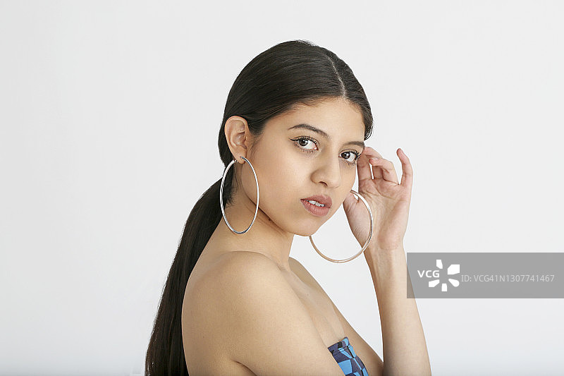 在影棚白色背景下手持马尾辫、身穿彩色连衣裙、佩戴大型银色耳环的拉丁裔千禧一代女性肖像图片素材