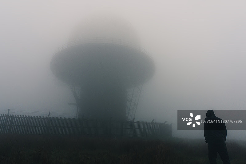 一个戴着头罩的人站在荒原上的雷达站旁的未来主义概念。在一个阴森、有雾的日子图片素材