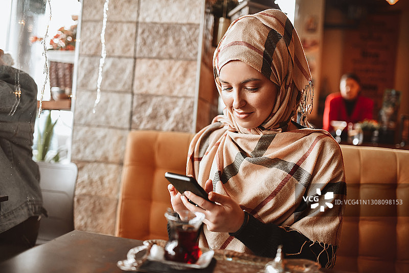 在餐厅使用智能手机的微笑穆斯林女性喝茶时光图片素材