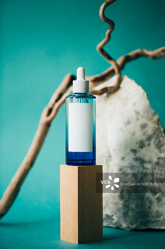 蓝色玻璃瓶精油或面部精华，木质底座，蓝绿色背景，植物枝条和粗糙石头，有机化妆品和天然成分概念，正面有复制空间图片素材