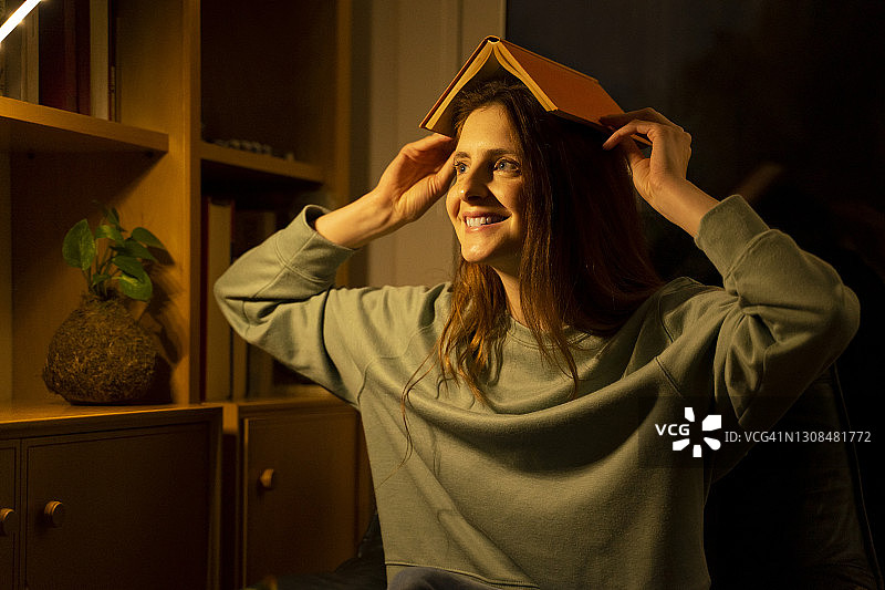 快乐的女人头顶着书，面带微笑，站在家里，目光移开图片素材