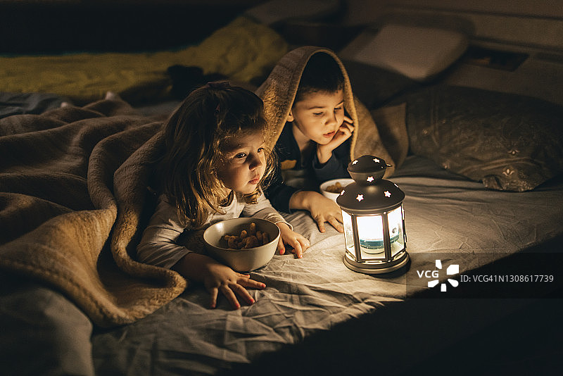 在黑暗房间里，盖着毯子，吃着零食，看着发光复古灯笼的兄妹图片素材