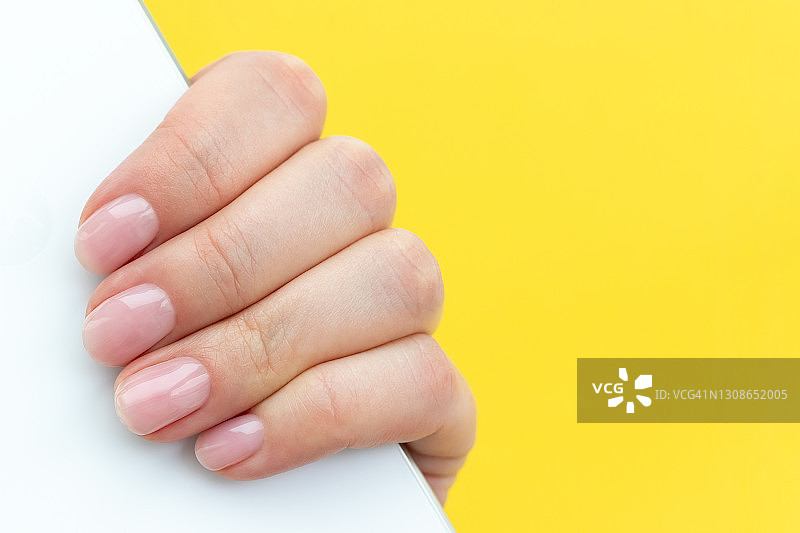 女人手部美甲特写：粉色裸色美甲，手持白色智能手机，黄色背景，含文案空间，选择性聚焦图片素材