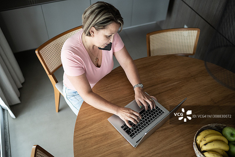 在家使用笔记本电脑并通过智能手机交谈的中年妇女图片素材