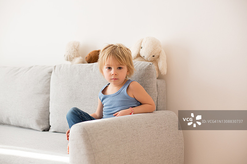 坐在阳光明媚的房间沙发上的金发小男孩，面带微笑图片素材