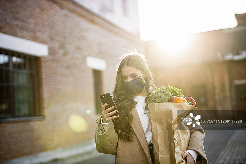 疫情期间戴黑色防护口罩，使用智能手机去购物的漂亮女人图片素材