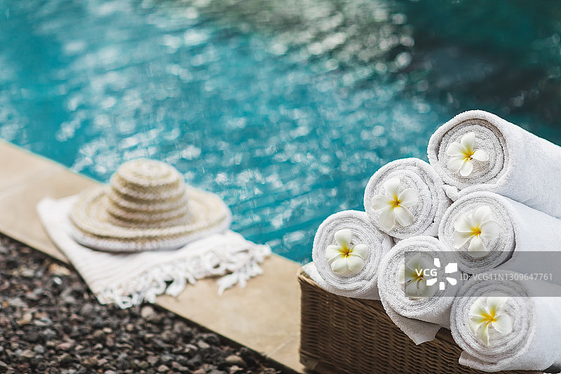 泳池边的白色卷沙滩巾、柳条箱和草帽及热带花卉图片素材