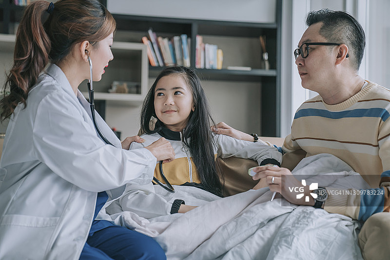 亚洲华人女医生上门拜访，检查小女孩的肺部呼吸，她那担心的父亲在她旁边的客厅里图片素材