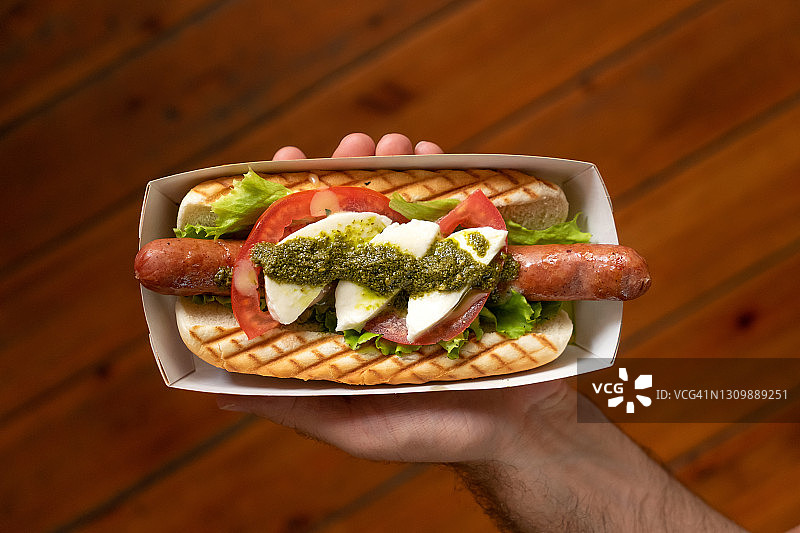 男性手中的热狗：烤面包夹热香肠、番茄和马苏里拉奶酪。外卖食品概念，营养快餐或午餐，顶视图图片素材