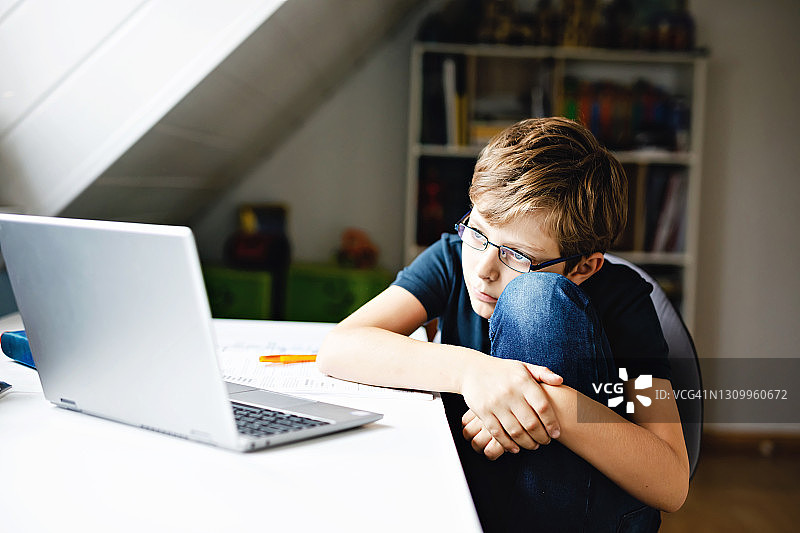 戴眼镜的男孩在家用笔记本电脑学习。可爱的孩子在做作业，使用笔记本和现代小工具。家庭学校教育概念。图片素材