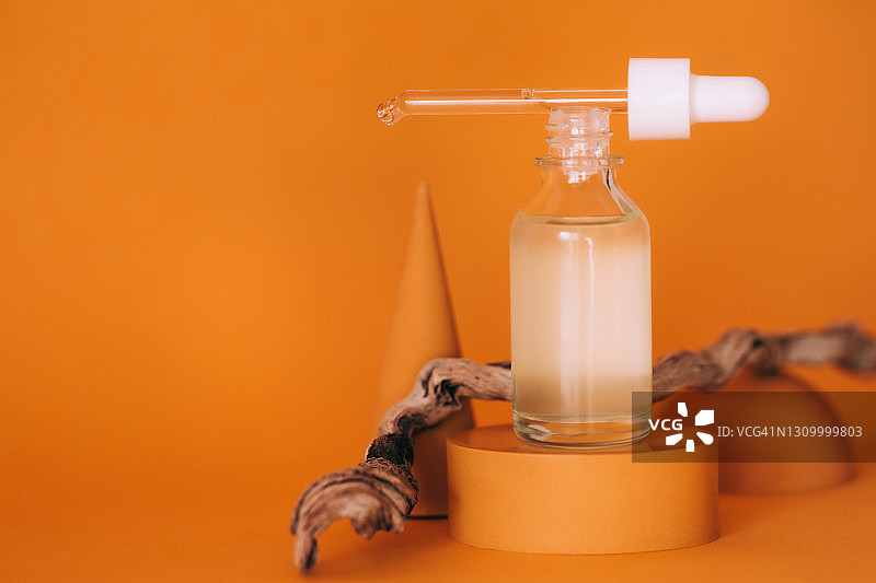 带有滴管的透明玻璃瓶和面部精华液，放置在混凝土橙色台上，靠近干燥的树枝、半球和锥体，位于明亮的橙黄色背景上，2021年潮流亮色，带有复制空间的前视图图片素材