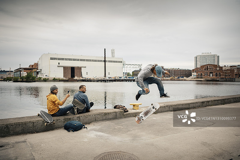 男子滑冰，男性朋友坐在运河旁，用智能手机对着天空拍照图片素材