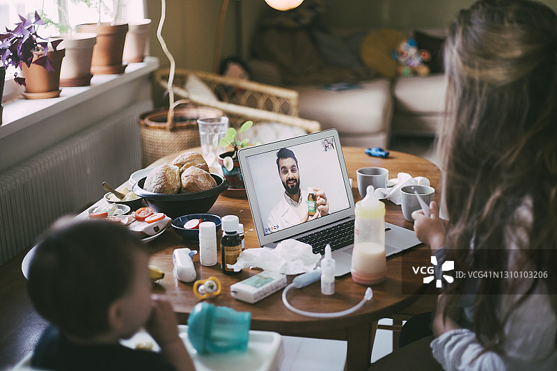 疫情期间母亲在客厅通过笔记本电脑视频通话向男医生咨询图片素材