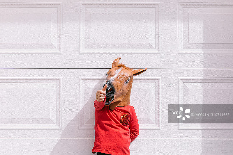 在白色车库门前，戴着马头乳胶面具并竖起拇指的小男孩图片素材