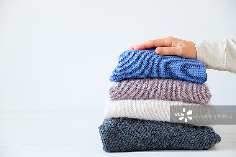 女人的手放在白色木桌上，堆叠着不同颜色的针织品。冬季和秋季温暖舒适的毛衣用于慈善事业。手工产品的存储、护理和洗涤概念。文本空间的副本。图片素材