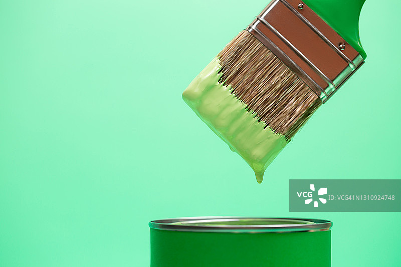 绿色的油漆从画笔刷毛滴入带有颜料的罐中，背景为柔和的翠绿色。家居维修概念。正面和特写图片素材