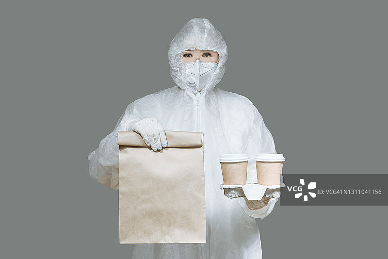 身穿防护服、戴口罩和手套的女人在灰色背景下手提纸袋和咖啡杯。食品配送服务图片素材