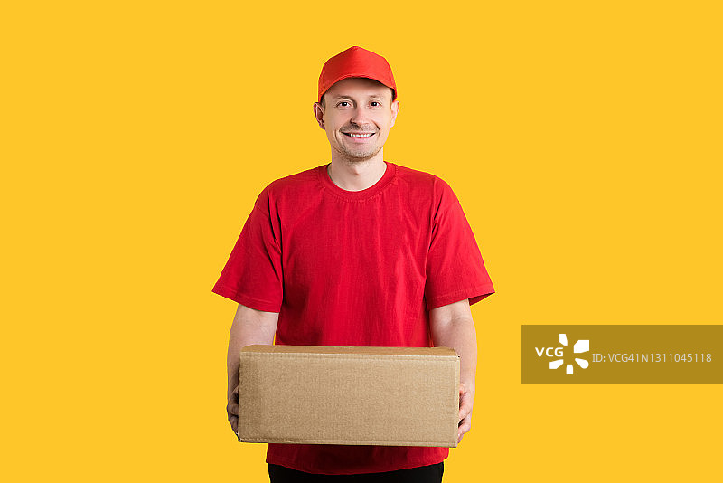 快递送货上门：微笑的男快递员拿着纸箱站在黄色背景前，网上购物概念图片素材