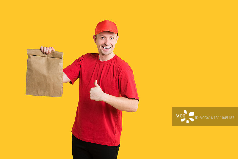 食品配送：红色制服快递员拿着环保包装在黄色背景上竖起大拇指图片素材