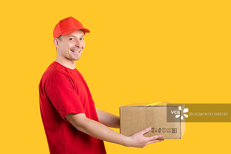 网店商品配送。快乐的男快递员身穿红色制服，在黄色孤立背景的工作室里拿着纸箱。留白图片素材