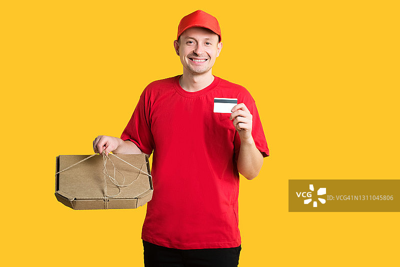 披萨外卖服务：穿红色制服的外卖员拿着盒子和信用卡，黄色背景隔离。留白图片素材