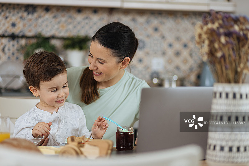年轻女子在桌旁使用笔记本电脑，她的幼子正在吃早餐图片素材