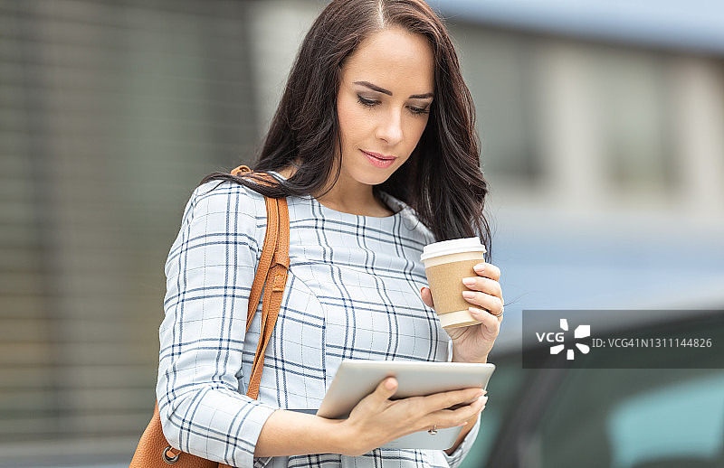 一位漂亮的女士在巴士站等候有轨电车时，阅读平板电脑并啜饮外卖咖啡。图片素材