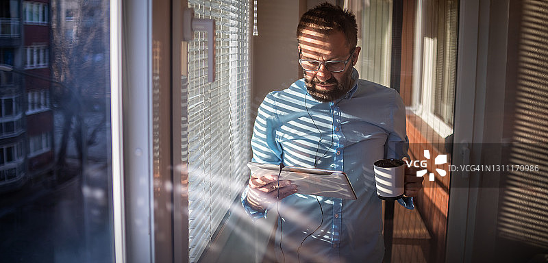 在数字平板电脑上工作的商人站在窗边，阳光在他身上投下真实的阴影。城市景观，客厅作为办公室 - 商人喝咖啡图片素材