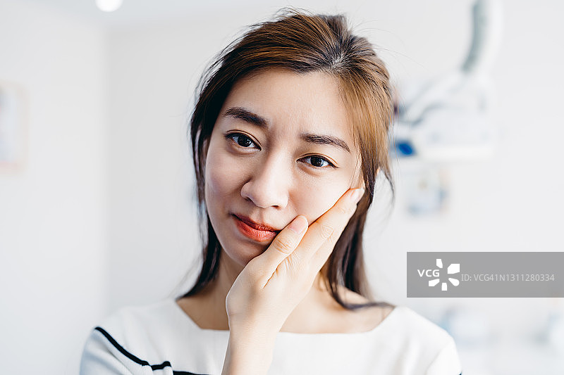 亚洲年轻女人在牙科诊所捂着脸，感到疼痛，牙痛概念图片素材