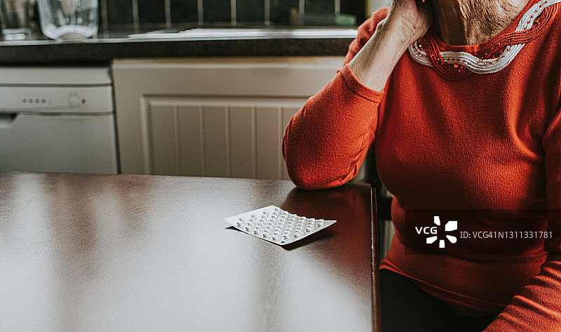 老人手扶着头坐在桌旁，看着一板药片图片素材