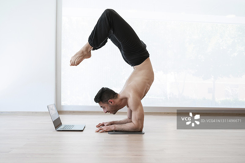 年轻的运动男子在家做瑜伽倒立并使用笔记本电脑图片素材