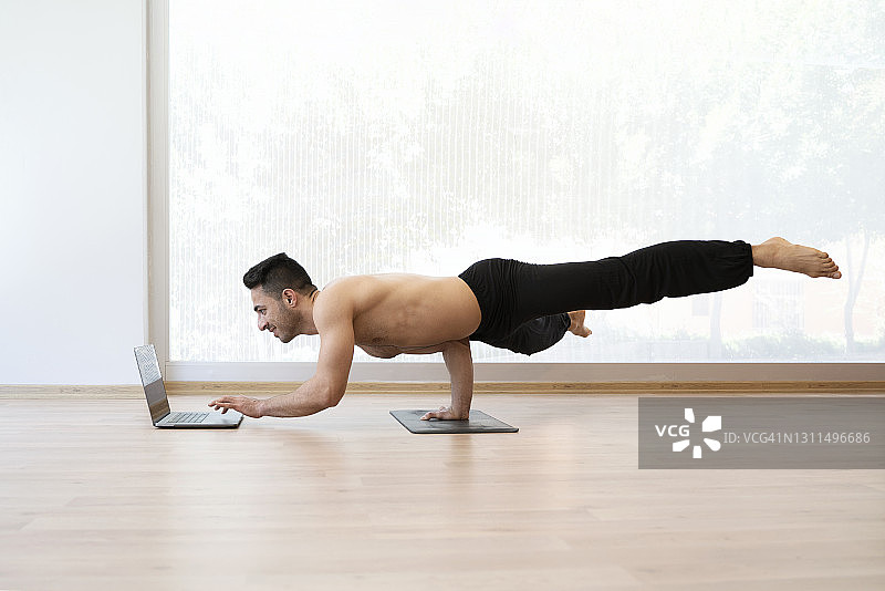 年轻的运动男子在家做瑜伽倒立并使用笔记本电脑图片素材