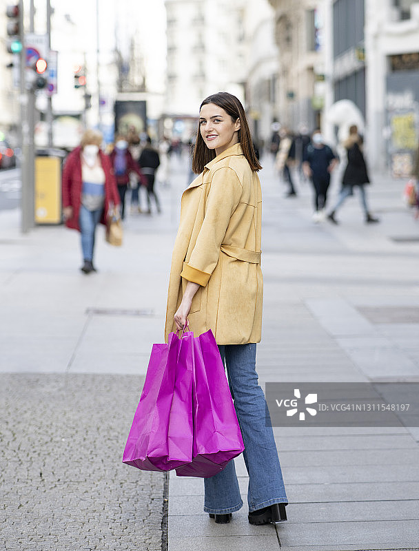 拎着洋红色购物袋在城市人行道上行走微笑的年轻女人图片素材